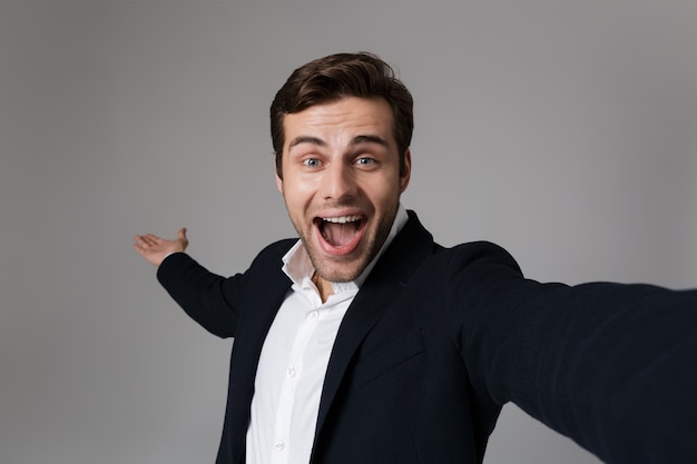 Image of young businessman 30s in formal suit screaming  while taking selfie photo, isolated over gray wall