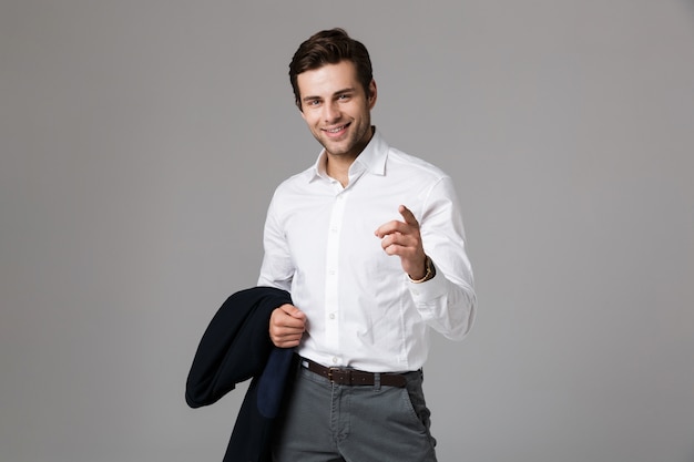 Image of young businessman 30s holding jacket and pointing finger, isolated over gray wall