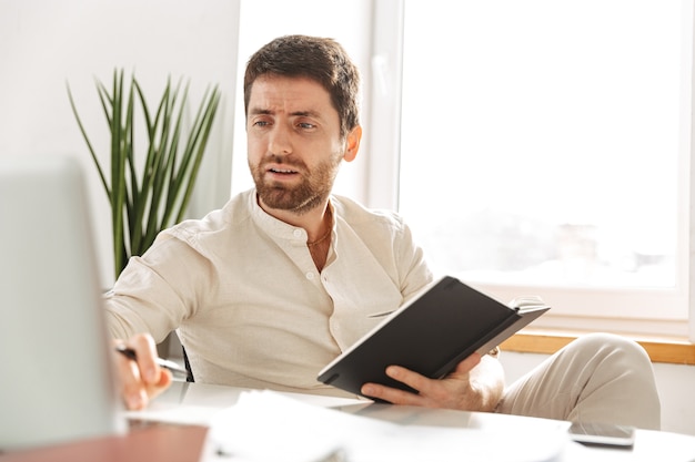 Image of young businessman 30s wearing white shirt using laptop and notebook, while sitting at table in modern workplace