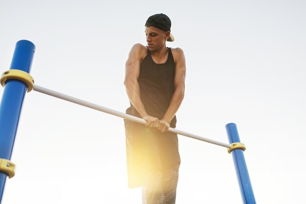 Image of young Caucasian muscular male doing pull ups exercises on horizontal bar outdoors Athletic runner training hard at sunny afternoon outside on the bar Sportsman working out against the sky