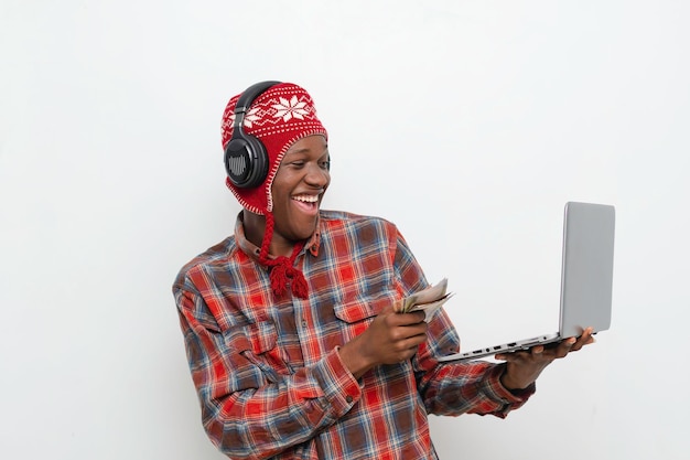 Изображение молодого веселого чернокожего мужчины в наушниках, обналичивающего иностранную валюту, держа в руках ноутбук