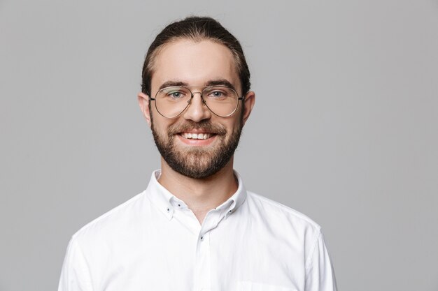 Image of a young cheerful happy handsome bearded man posing isolated over grey wall wearing glasses.