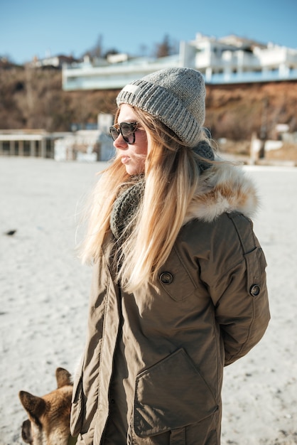 Photo image of young concentrated woman wearing sunglasses walks in winter beach with dog on a leash.