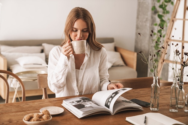 コーヒーを飲みながら雑誌を読んでオフィスで屋内に座っている若いかわいい美しい女性の画像。