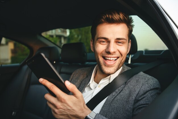 Image of young director man in suit holding smartphone and smiling, while back sitting in business class car with safety belt