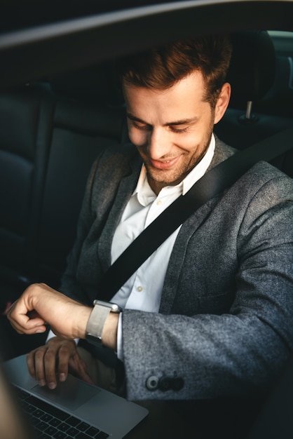 Image of young director man in suit working on laptop and looking at wrist watch, while back sitting in business class car with safety belt