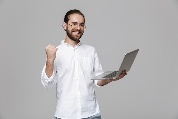 ラップトップコンピューターを使用して眼鏡をかけている灰色の壁の上に孤立してポーズをとっている若い感情的な幸せな楽観的なハンサムなひげを生やした男の画像は、勝者のジェスチャーを作ります。