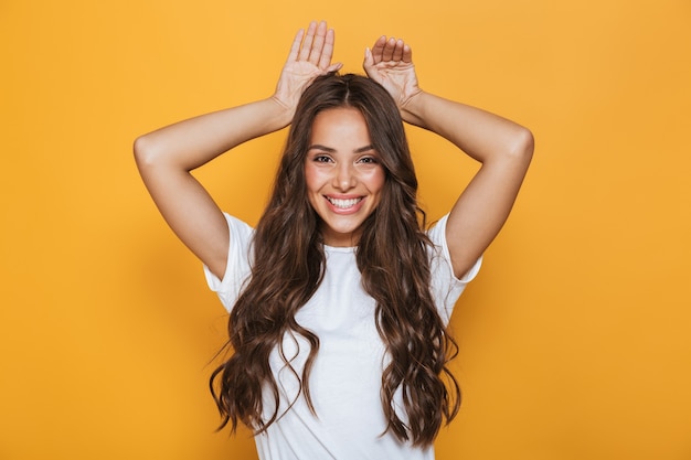 Image of young european woman 20s with long hair smiling and showing rabbit ears at her head, isolated over yellow wall