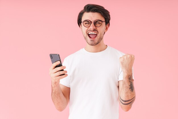 Image of young excited man wearing eyeglasses holding cellphone and showing winner gesture isolated