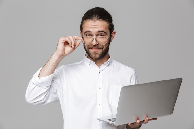 Image of a young handsome bearded man posing isolated over grey wall wearing glasses using laptop computer.