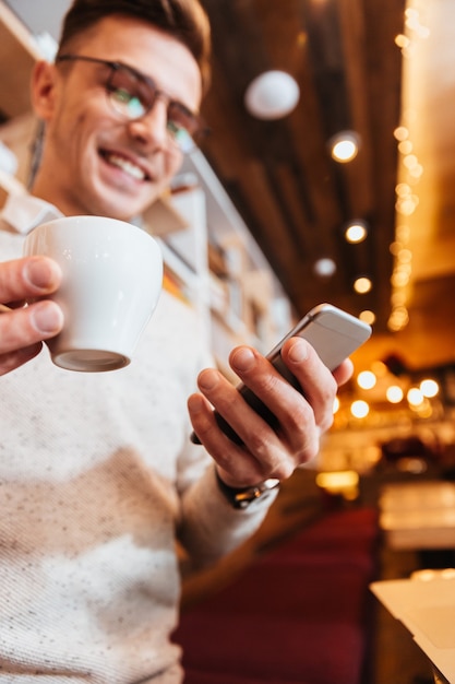 携帯電話を使用しながらカフェに座って眼鏡をかけている若い幸せな集中男性の画像。