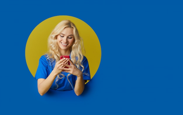 Image of young happy woman standing over a blue wall. woman peep out of a hole in the wall. Looking to the side to take a selfie on the mobile phone.