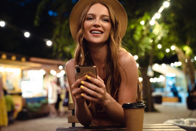 携帯電話を使用して夕方の夜に屋外のカフェに座っている若い笑うポジティブな女性の画像。