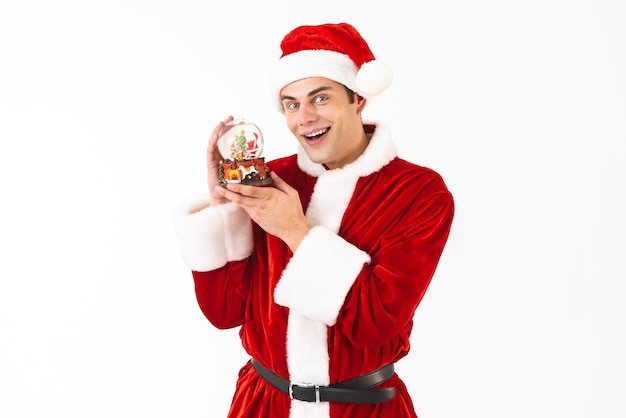 Image of young man 30s in santa claus costume and red hat holding christmas ball