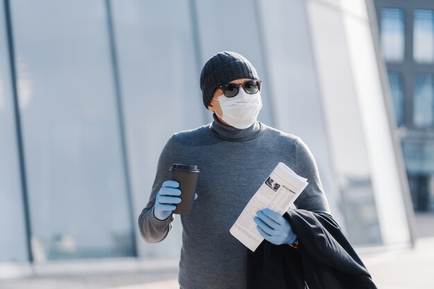 Image of young man wears sterile mask and rubber medical gloves, looks aside, walks through city during infectious disease spread, drinks coffee, folded newspaper. Coronavirus prevention concept