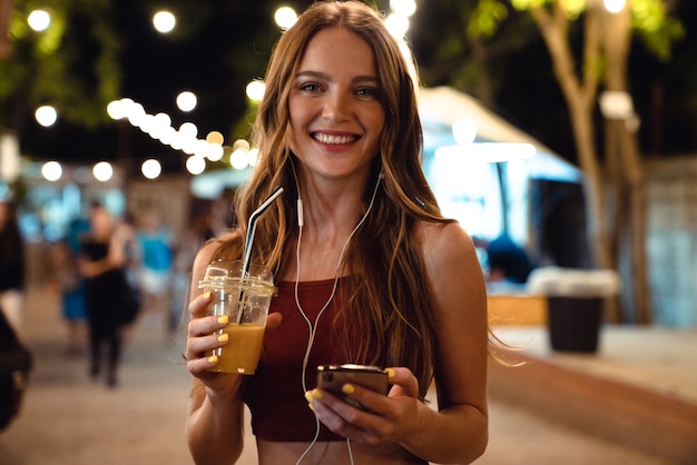 Immagine di una giovane donna felice ottimista che cammina per strada all'aperto bevendo succo la sera sera usando il telefono cellulare.