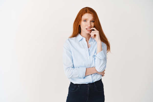 Image of young redhead female office worker biting finger and looking indecisive having doubts or worries standing over white background