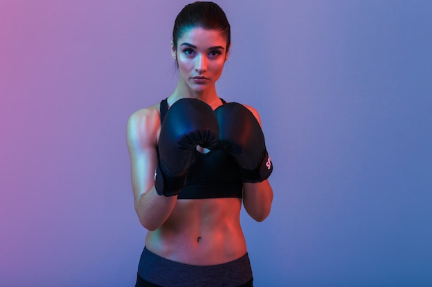 Image of young sports woman in tracksuit and black boxing gloves fighting in gym, isolated over purple wall