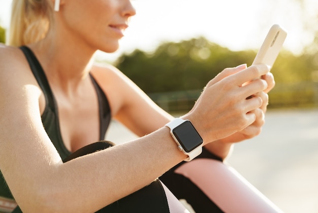 Image of young sportswoman wearing tracksuit and earpods holding cellphone while doing workout outdoors