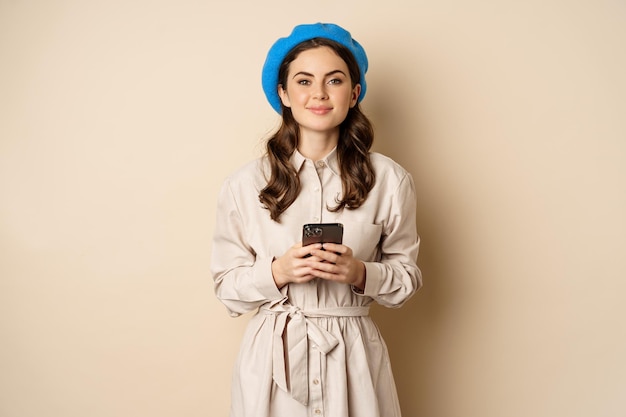 Image of young stylish french woman in trench coat standing with smartphone using mobile phone app a...