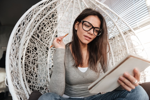 Image of young thoughtful lady wearing eyeglasses sitting on chair indoors while writing notes in notebook. Look at notebook.