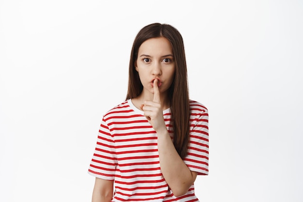 Image of young woman asking to be quiet, keep secret, hushing with finger on lips and frowning, stay silent, taboo sign, standing against white background.