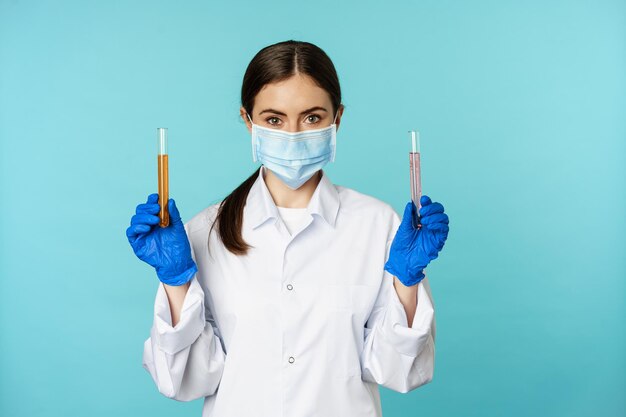 Image of young woman doctor lab worker doing research holding test tubes wearing medical face mask and rubber gloves blue background person