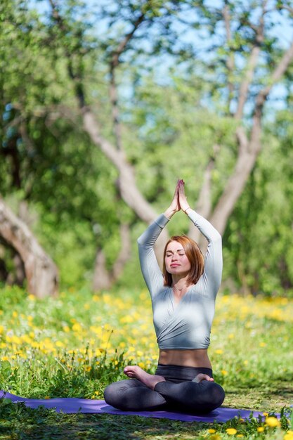 Immagine di una giovane donna che fa yoga con le braccia alzate seduta nella posizione del loto su un tappeto blu nella foresta il giorno d'estate