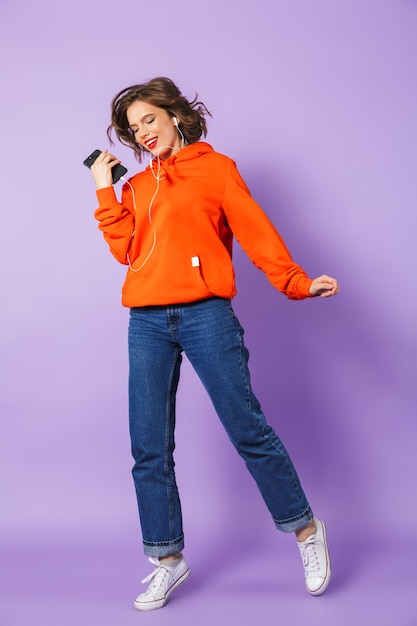 Image of young woman isolated over purple wall wall listening music with earphones holding phone.