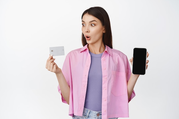 Image of young woman looking at credit card with excitement showing smartphone screen standing over white background Copy space
