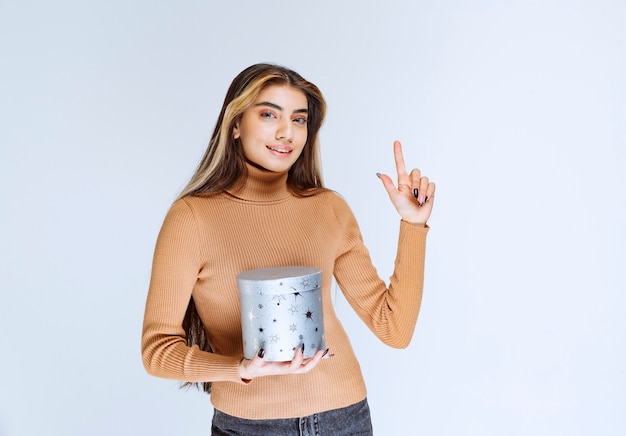 Image of a young woman model in brown sweater with a gift box pointing up.