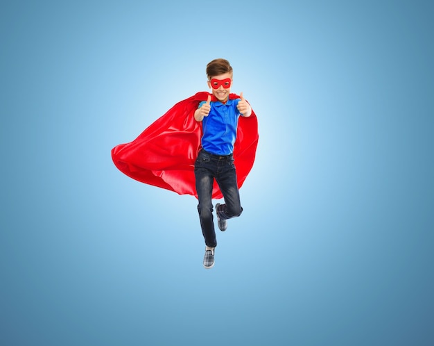 imagination, gesture, childhood, movement and people concept - boy in red super hero cape and mask flying in air and showing thumbs up over blue background