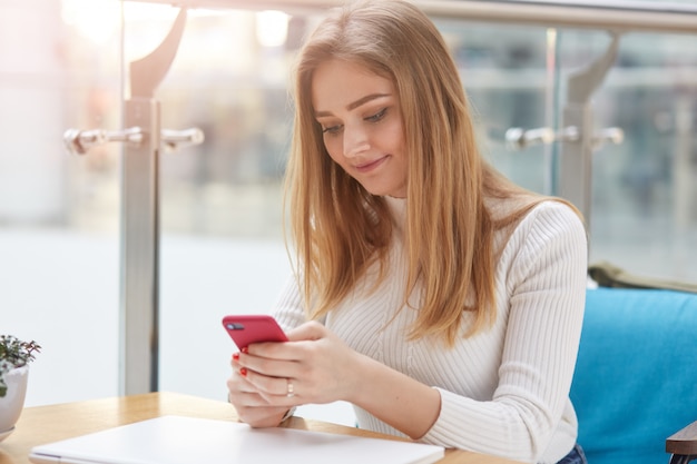 Immagina un'affascinante donna vestita di una maglietta bianca casual, ha lunghi capelli biondi, seduta al mattino al ristorante, in possesso di telefono, controllando e-mail o social network. concetto di persone e tecnologia.