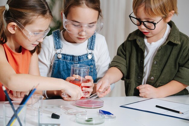 Immersed little kids doing home science project, filling glass dish. All behid table, wearing glasses. Chemical glassware and colored liquids on the table.