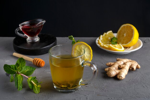 Immune gemberdrank met honingmunt en citroen in een glazen beker in de buurt van ingrediënten op een betonnen achtergrond.