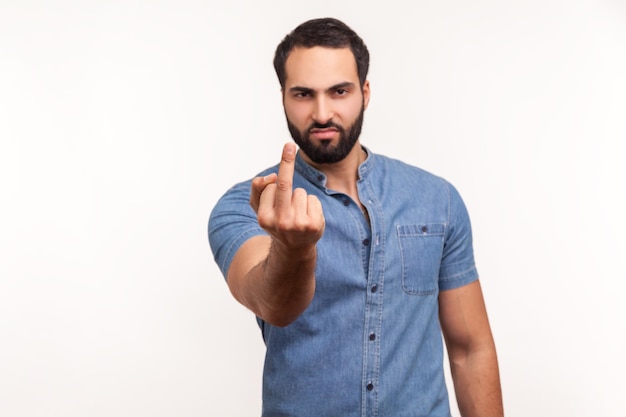 Невежливый агрессивный мужчина показывает средний палец и просит прекратить смотреть в камеру с негативом, неуважительным поведением. Крытая студия снята на белом фоне