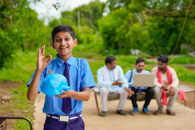 importance of saving concept : Smart indian little boy standing and holding piggy bank in hand at home.