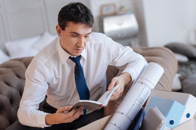 Important notebook. Calm concentrated young man looking at the notes in his notebook while sitting in front of a box with personal things and reading important information