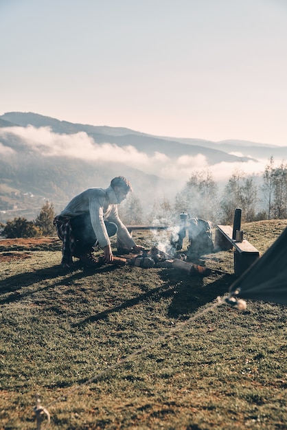 Important part of camping. Young man making a campfire while sitting near the tent in the mountains