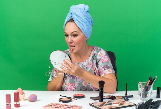 Impressed adult caucasian woman with wrapped hair in towel sitting at table with makeup tools applying lip gloss holding and looking at mirror isolated on green wall with copy space