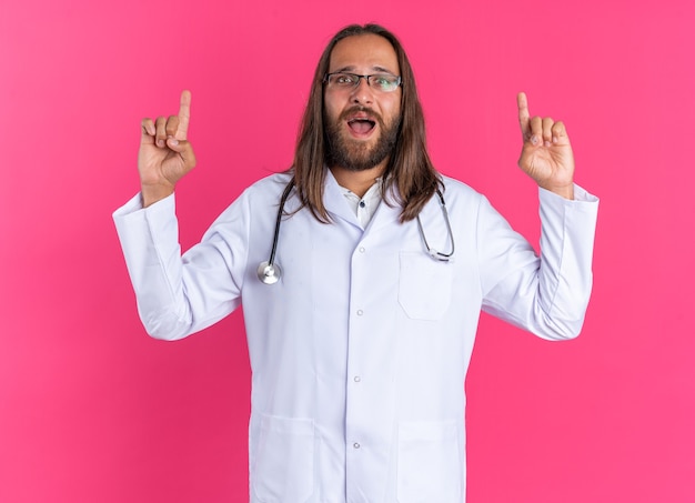 Impressed adult male doctor wearing medical robe and stethoscope with glasses pointing up 