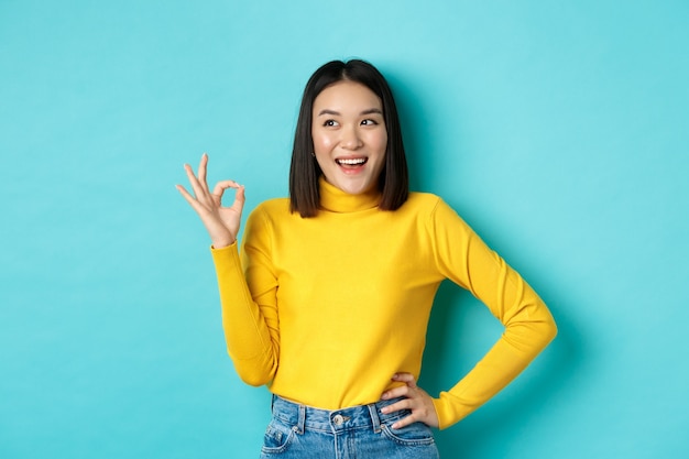 Impressed asian woman in yellow sweater, looking left amazed and showing OK sign in approval