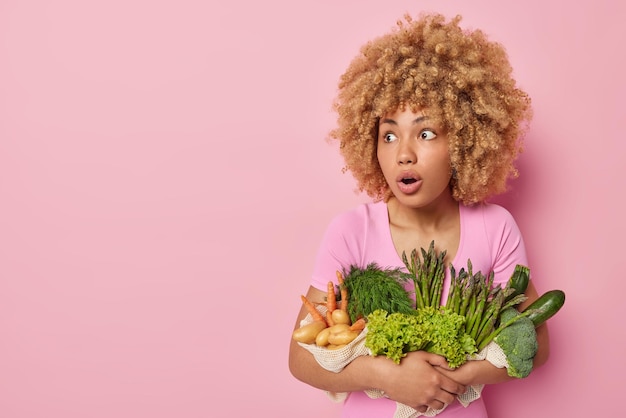 Impressed astonished curly haired woman embraces green grocery fresh vegetables grown in garden eats only organic food looks amazed away isolated over pink background blank space for promotion