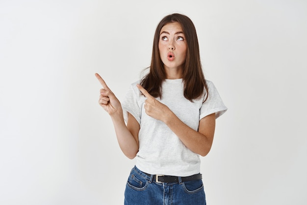 Impressed brunette girl checking out promo sale, pointing and staring at upper left corner advertisement, standing over white wall