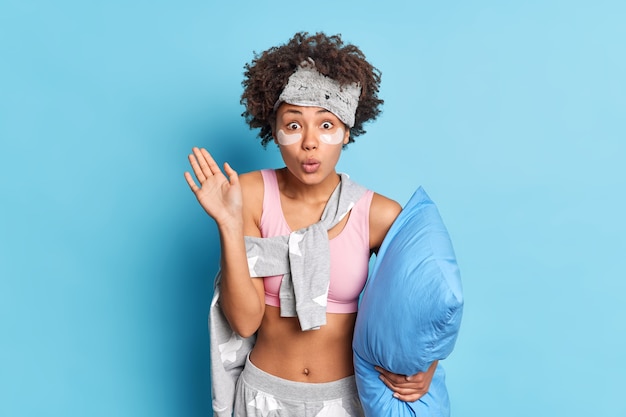 Impressed curly haired woman looks with wondered expression stares surprisingly at front keeps hand raised dressed in pajama poses with pillow isolated over blue wal