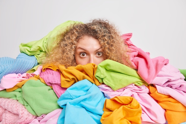 Impressed curly haired young European woman hidden behind pile of multicolored laundry going to do washing at home isolated over white 