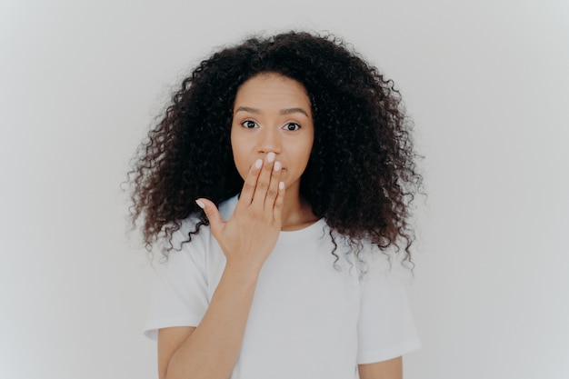 Impressed curly young woman covers mouth with palm tries to be speechless