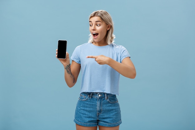 Impressed enthusiastic good-looking female blonde with cool tattoo on arm dropping jaw from thrill and excitement pointing at device screen over blue wall