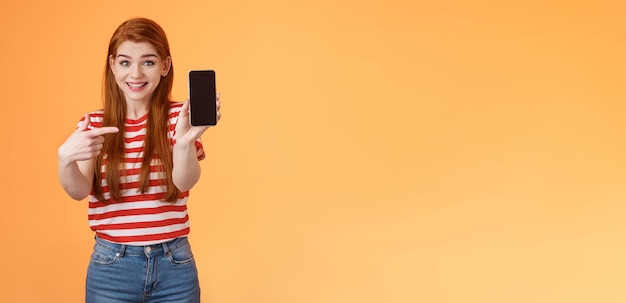 Photo impressed excited happy redhead woman smiling broadly hold smartphone pointing cellphone screen thri