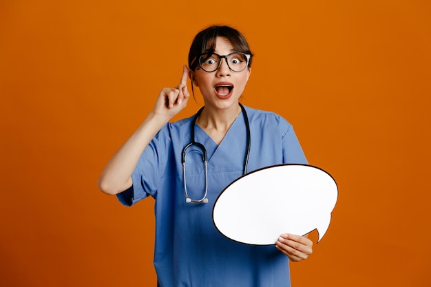 Impressed holding speech bubble young female doctor wearing uniform fith stethoscope isolated on orange background